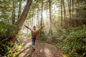 a women with open arms walking in a forest with rays of sunlight flowing through forest