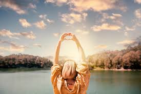 picture of a person with their arms stretches above their head looking toward body of water and sky with clouds
